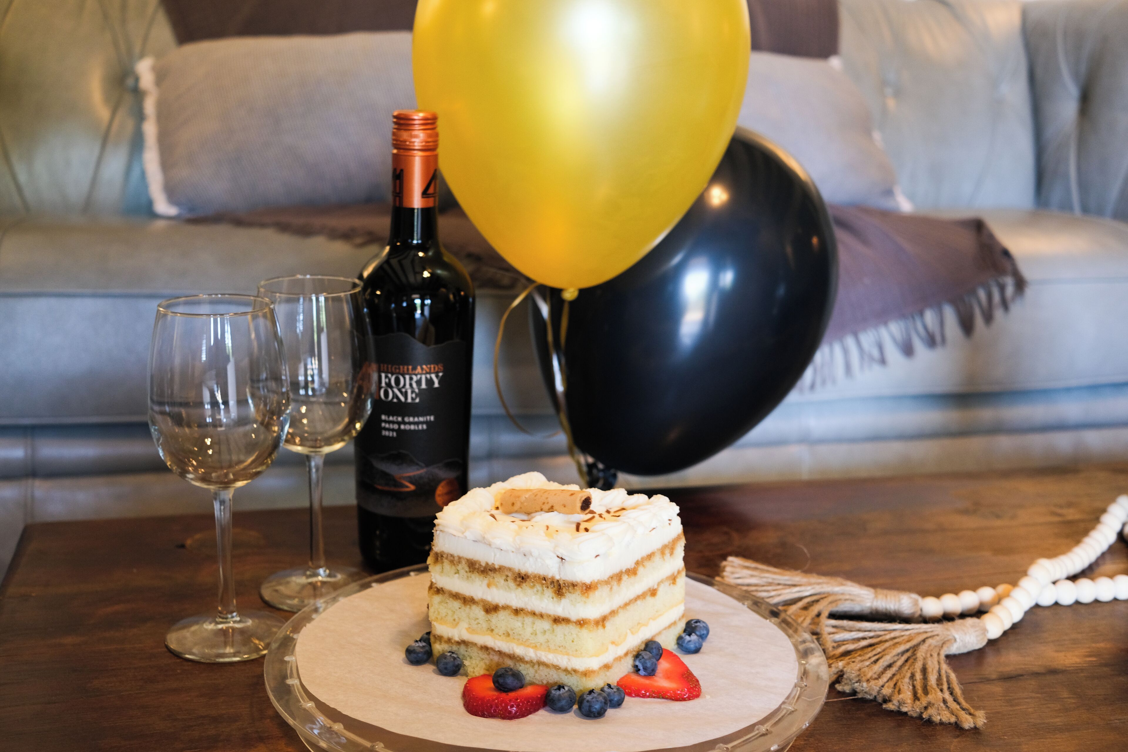 A slice of cake on a glass plate with strawberries and blueberries, a bottle of wine, and two wine glasses on a wooden table.