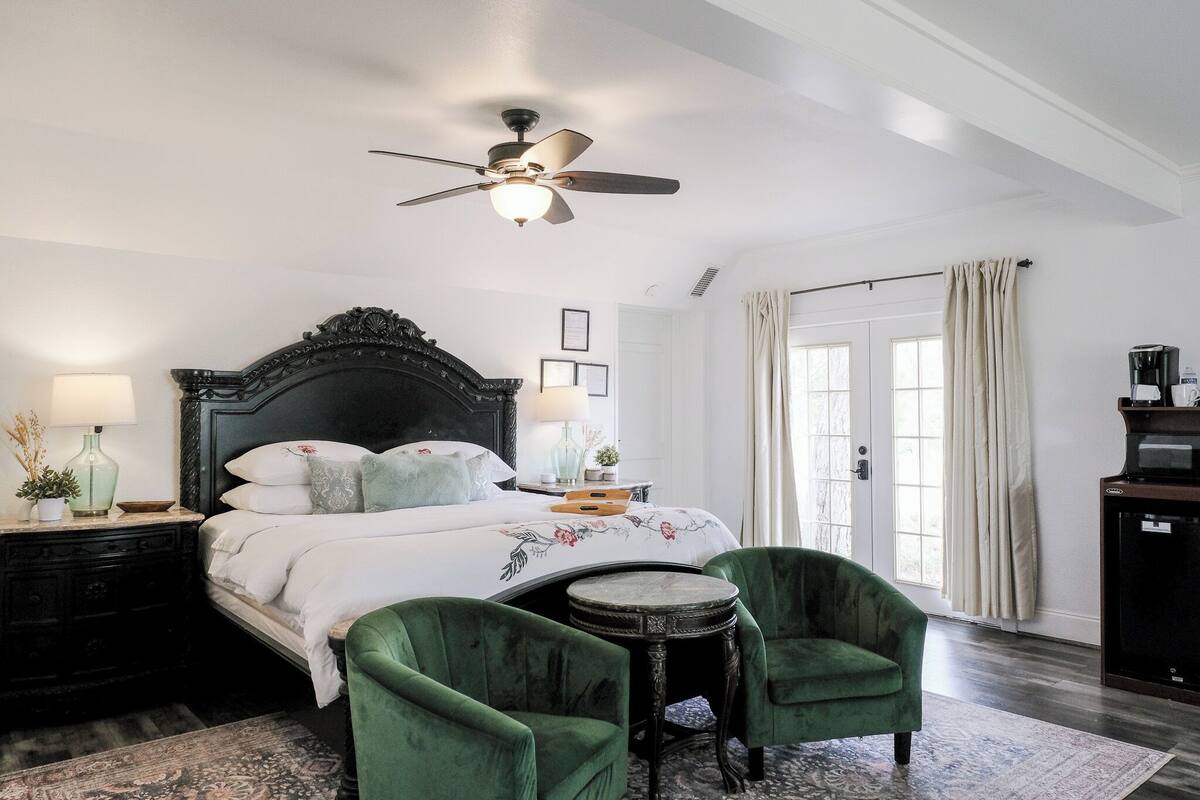 A bedroom with a large bed, a ceiling fan, a small table, two green armchairs, a nightstand, a lamp, a potted plant, and a window with curtains.