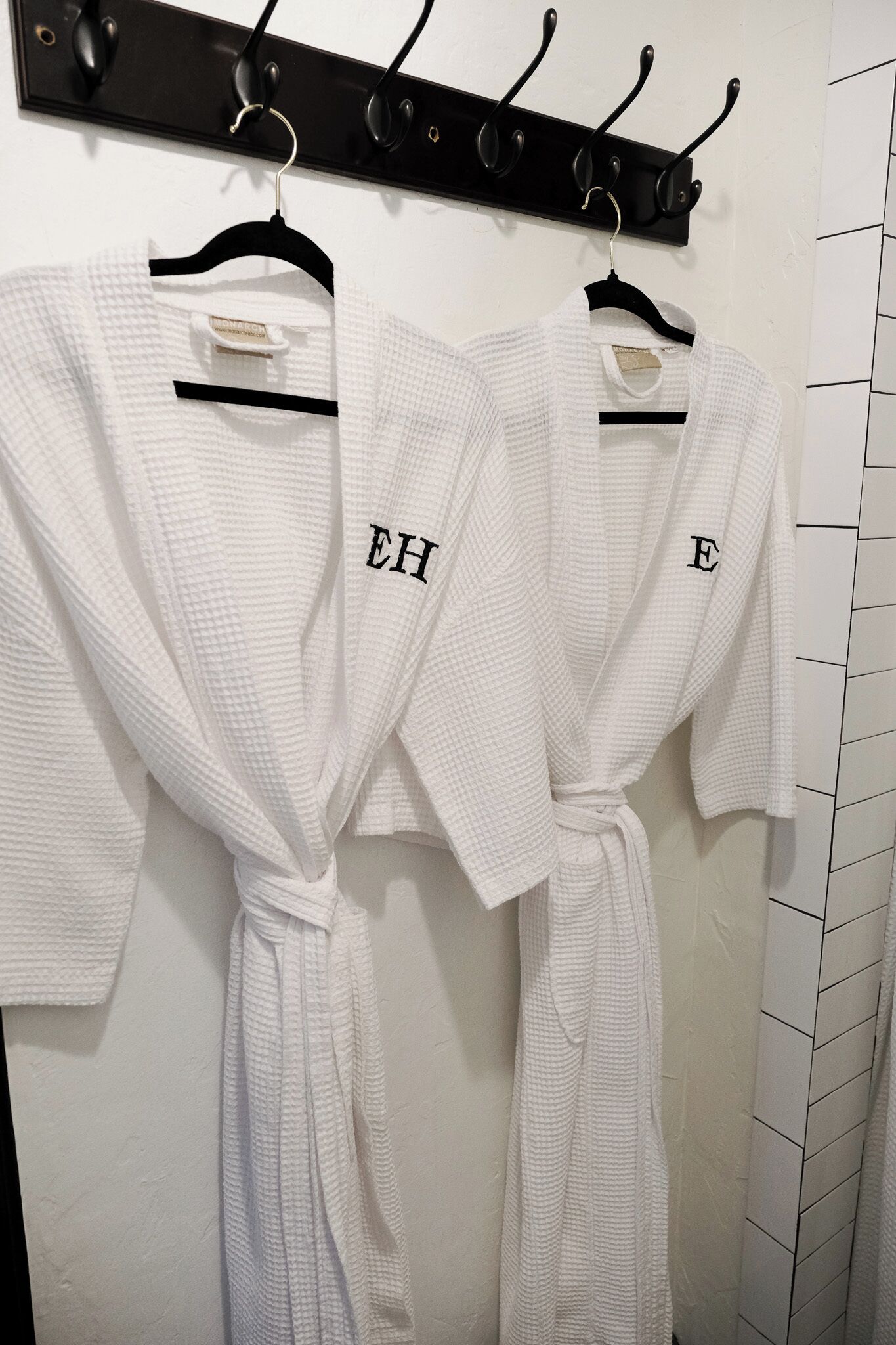 Deux robes blanches sont suspendues à un porte-serviettes noir fixé au mur, dans une salle de bain.
