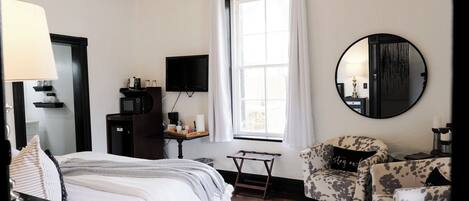 A bedroom with a bed, a desk, a chair, a TV, a window with curtains, and a mirror.