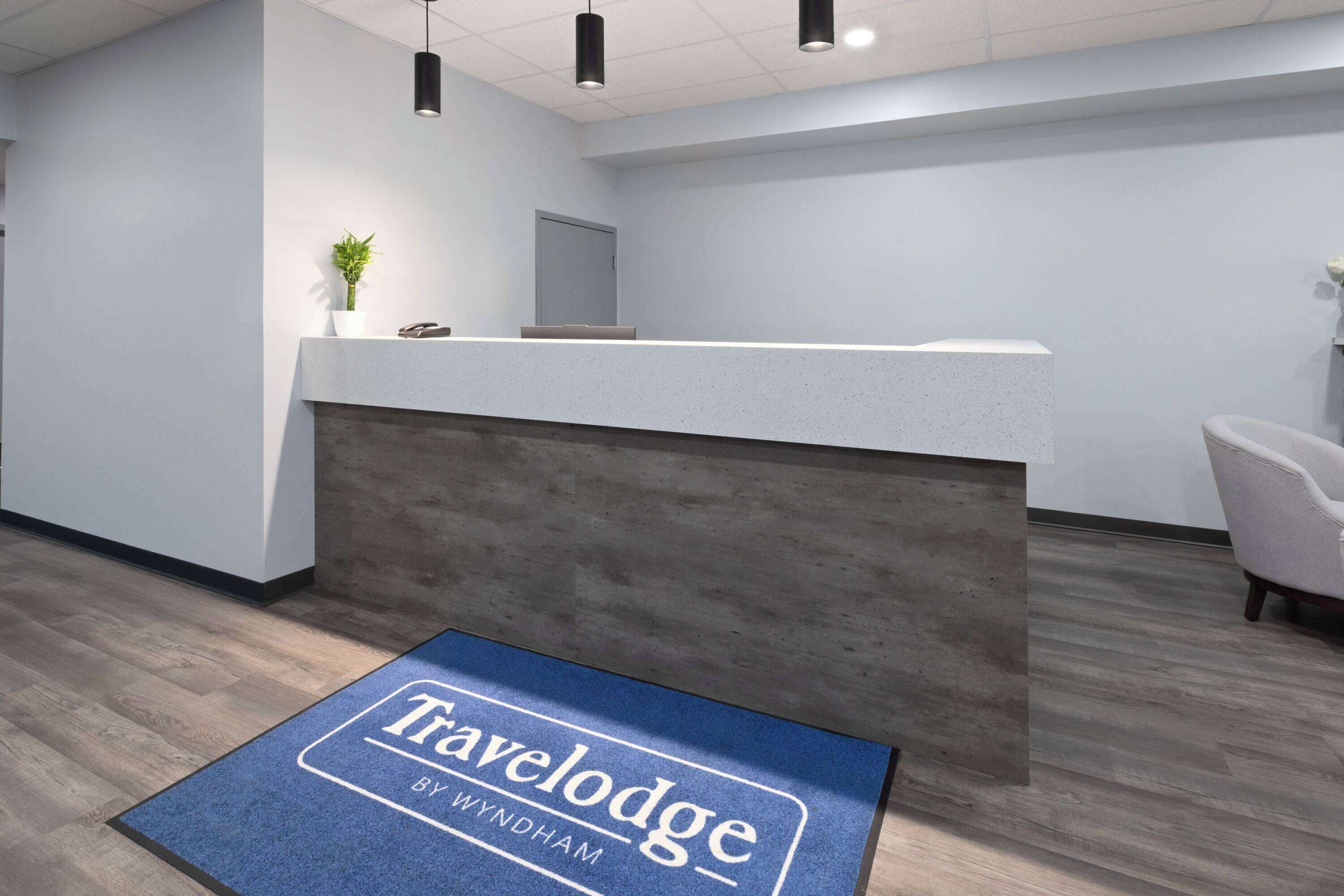 A reception area with a reception desk, a potted plant, a chair, and a blue mat with "Travelodge By Windham" text.