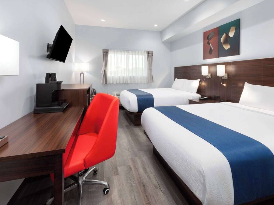 Une chambre d’hôtel avec deux lits, un bureau, une chaise rouge, un téléviseur à écran plat fixé au mur et une fenêtre avec des rideaux.