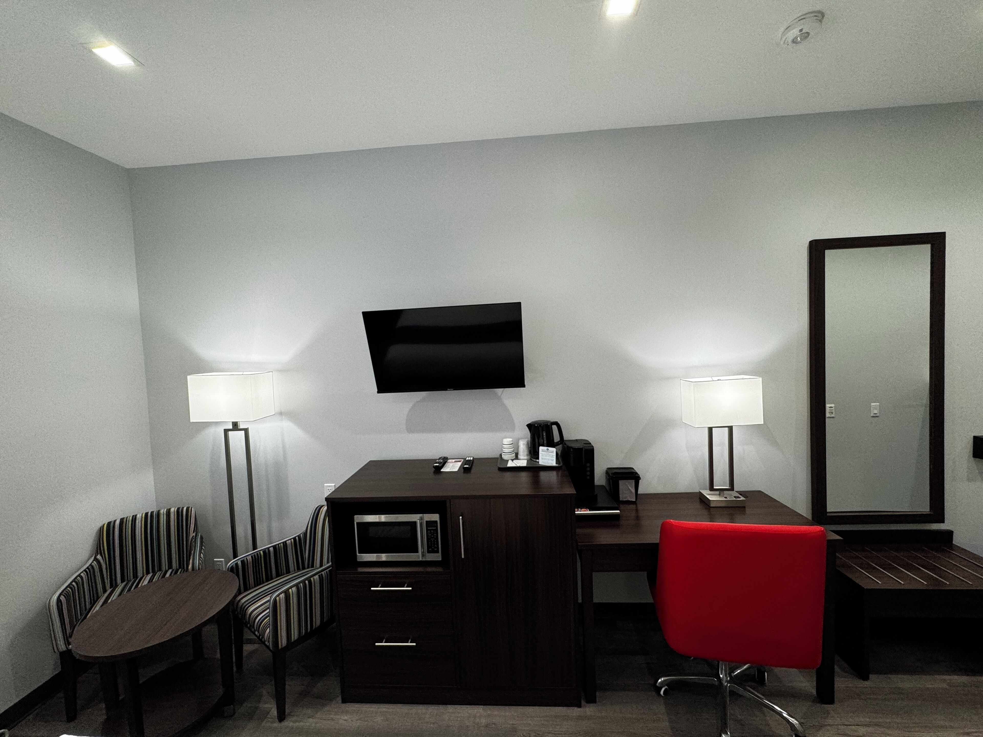 Habitación de hotel con un escritorio de madera oscura, una silla roja, un televisor montado en la pared y dos lámparas de pie.