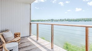 A wooden deck with a railing overlooking a calm lake, wicker furniture, and a white wall.