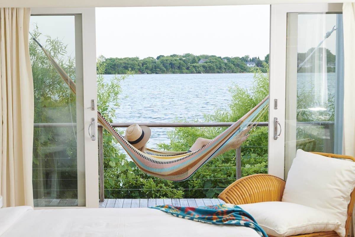 A bedroom with a large window, a hammock, a wicker chair, and a bed with white bedding.