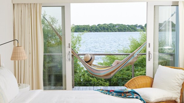 A bedroom with a large window, a hammock, a wicker chair, and a bed with white bedding.