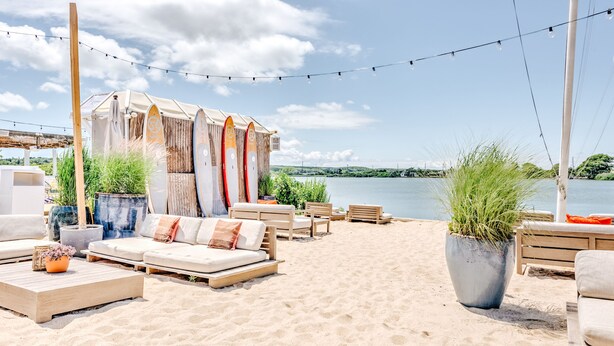 A beachside lounge with outdoor seating, a wooden pergola, and surfboards against a backdrop of a lake and clear sky. - The Surf Lodge (Montauk)
