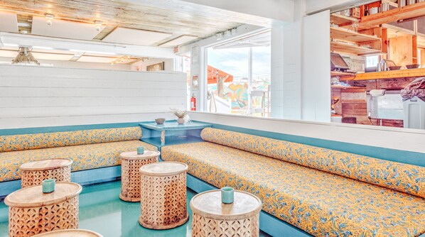 A nautical-themed seating area with patterned cushions, round stools, and a view of the sea through large windows.