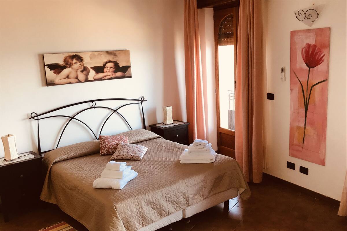 A neatly made bed with a quilted cover and pillows, a bedside table with a lamp, a painting on the wall, and a window with curtains.