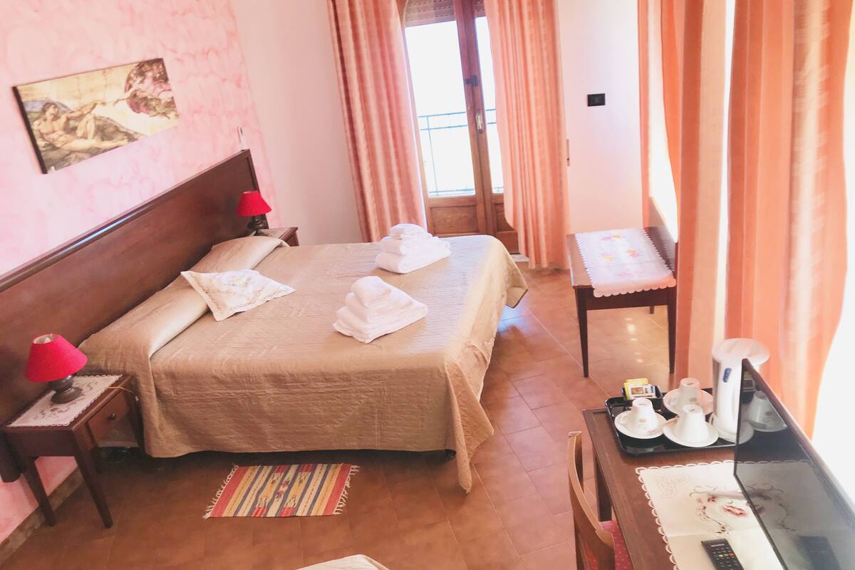 A hotel room with two beds, a wooden headboard, a red lamp, a small table with a remote control, and a window with curtains.