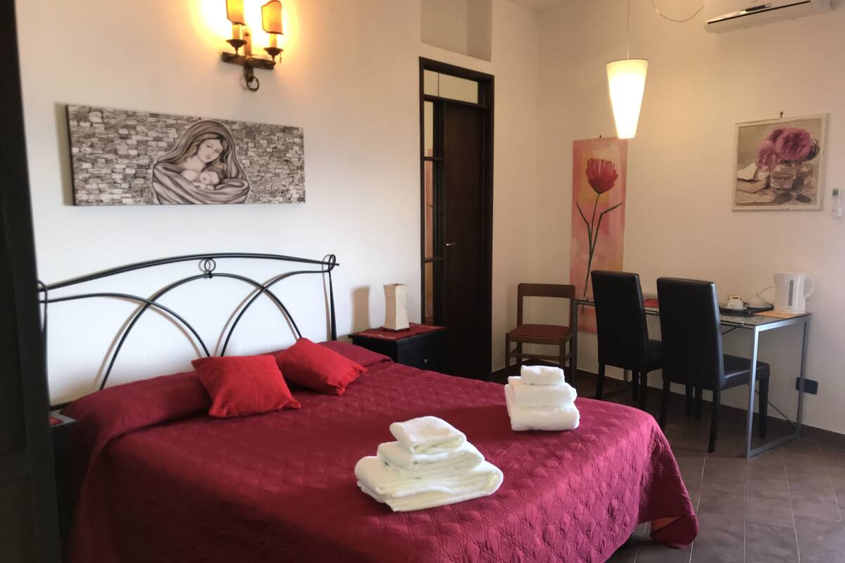 A neatly made bed with red bedding and white towels, a desk with chairs, and artwork on the walls.
