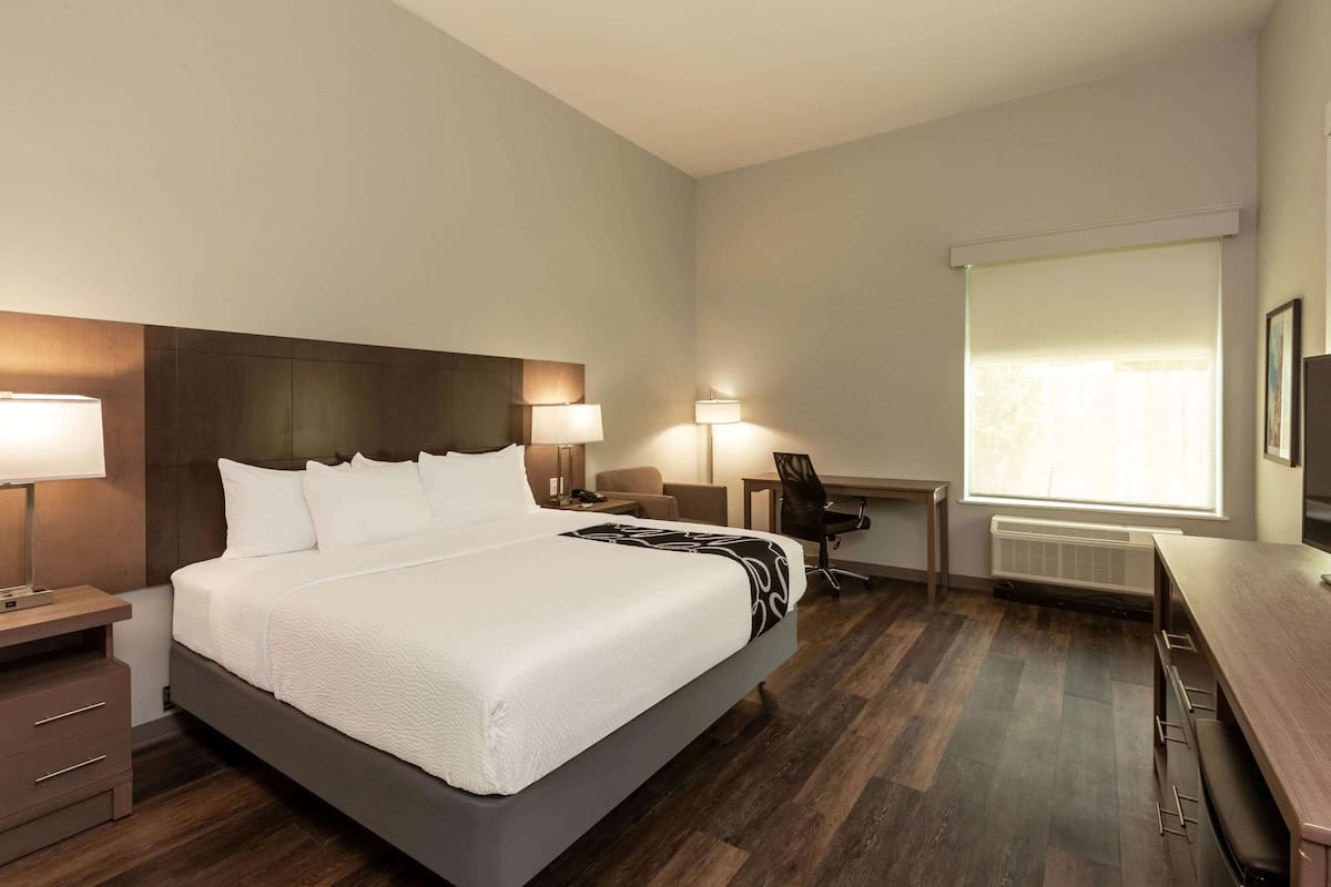 A hotel room with a large bed, bedside lamps, a desk, a chair, and a large window with blinds.