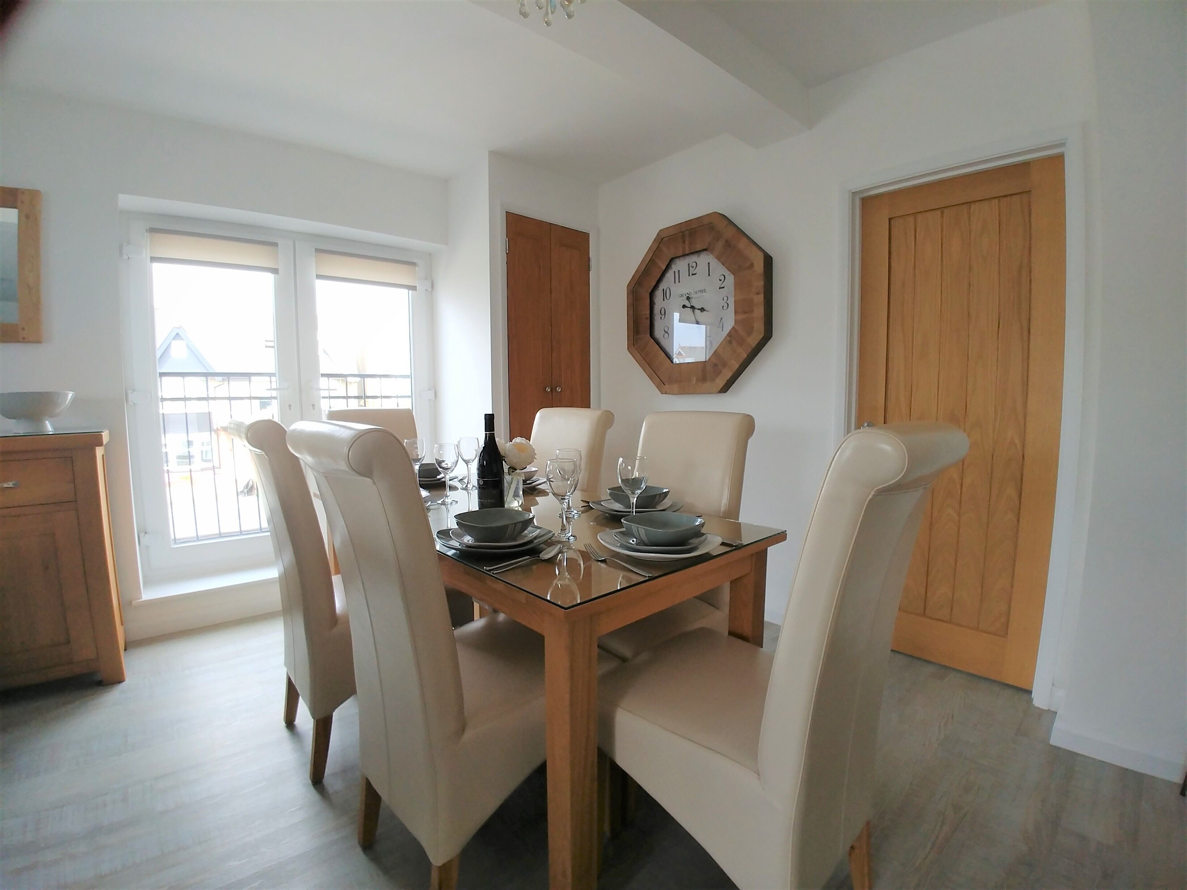 A dining area with a wooden table set for four, surrounded by beige chairs. A large octagonal clock is mounted on the wall above the table.