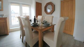 A dining area with a wooden table set for four, surrounded by beige chairs. A large octagonal clock is mounted on the wall above the table.