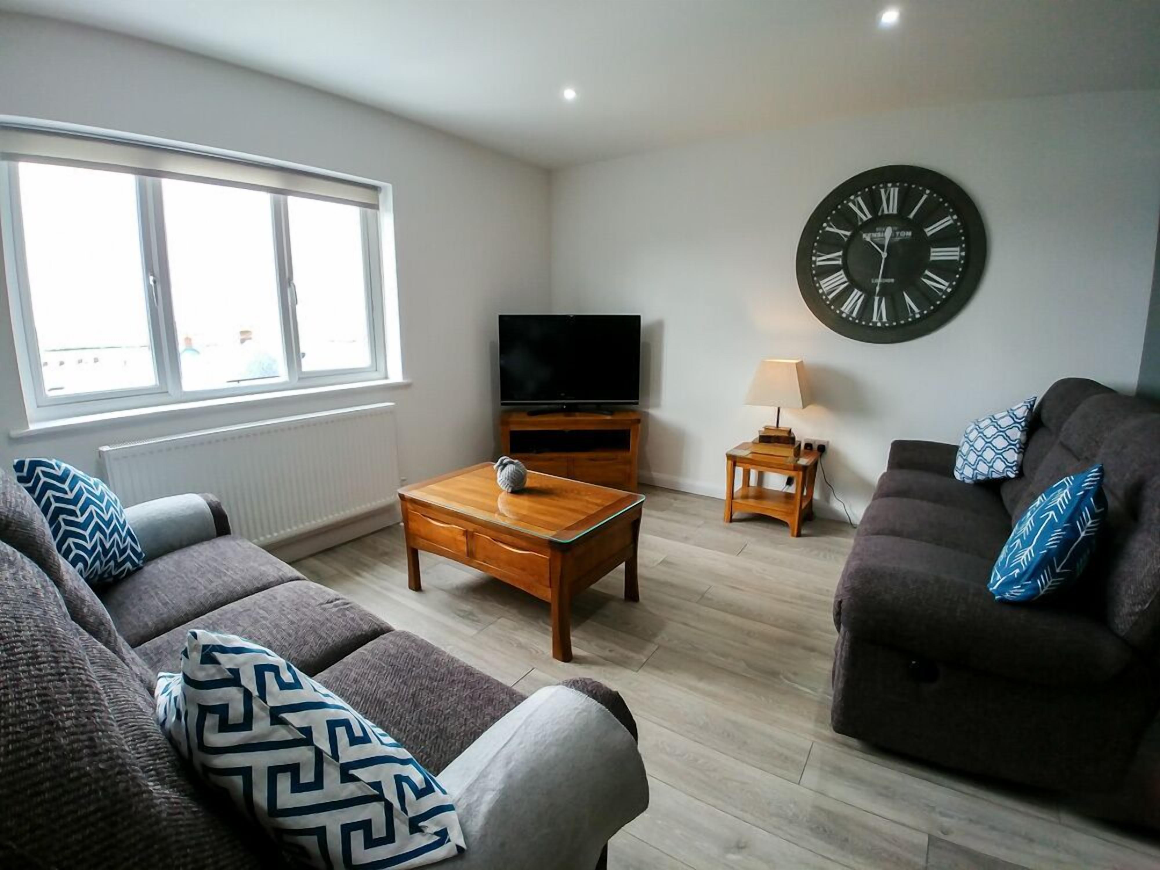 A living room with a sofa, a coffee table, a TV, a clock, and a window.