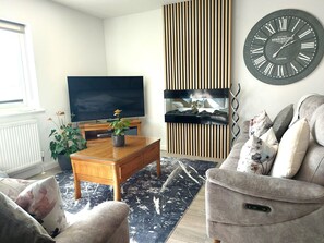 A living room with a flat-screen TV, a wooden coffee table, a grey sofa, and a decorative wall clock. - Meadow View Apartments (Newquay)
