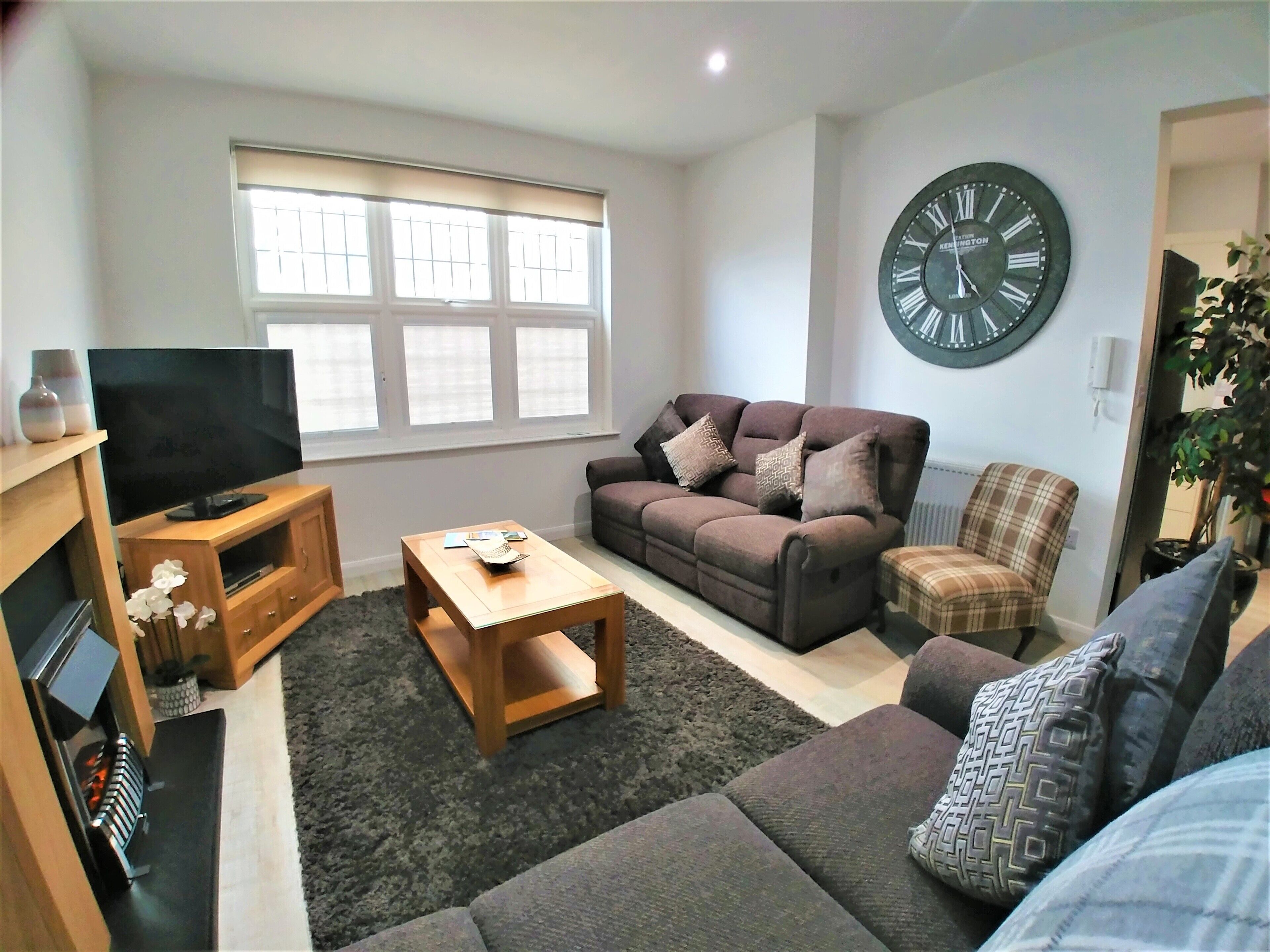 A living room with a sofa, a coffee table, a TV on a wooden stand, a large wall clock, and a window with blinds.