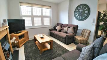 A living room with a sofa, a coffee table, a TV on a wooden stand, a large wall clock, and a window with blinds.