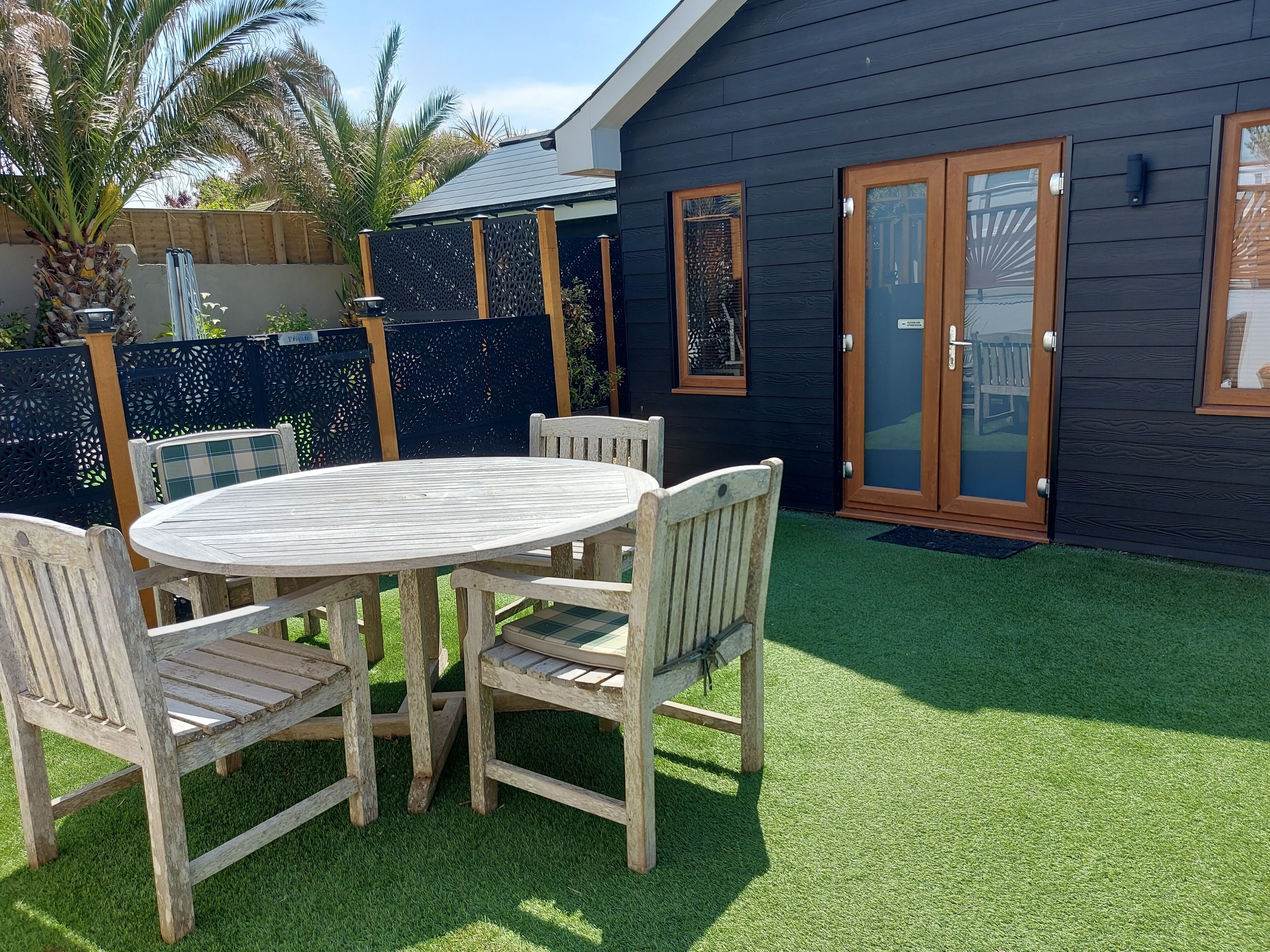 A wooden deck with a round table and chairs, a dark wooden building with a glass door, and a green artificial lawn.