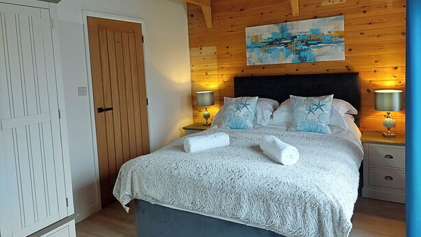 A bedroom with a wooden ceiling, a bed with a white and blue patterned bedspread, two bedside lamps, and a painting above the bed. - Meadow View Apartments (Newquay)