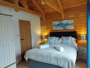 A bedroom with a wooden ceiling, a bed with a white and blue patterned bedspread, two bedside lamps, and a painting above the bed. - Meadow View Apartments (Newquay)