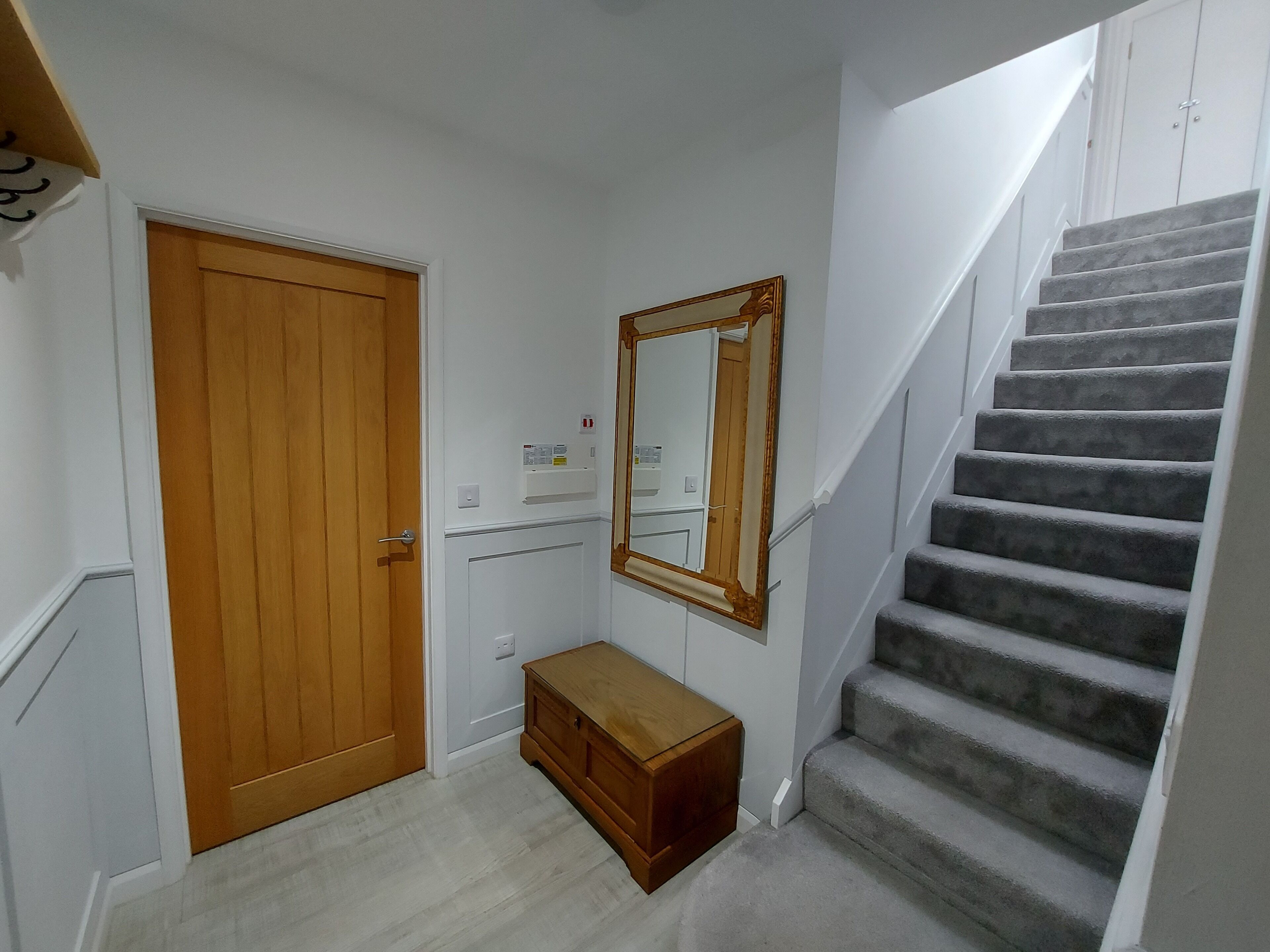 Un pasillo con una puerta de madera, un espejo, un banco y una escalera con alfombra.