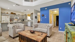 A spacious living area with a kitchen, two armchairs, a coffee table, and a television on a green cabinet. - Spacious 6 bed pool home with spa and games room in Emerald Island, sleeps 15 (Kissimmee)