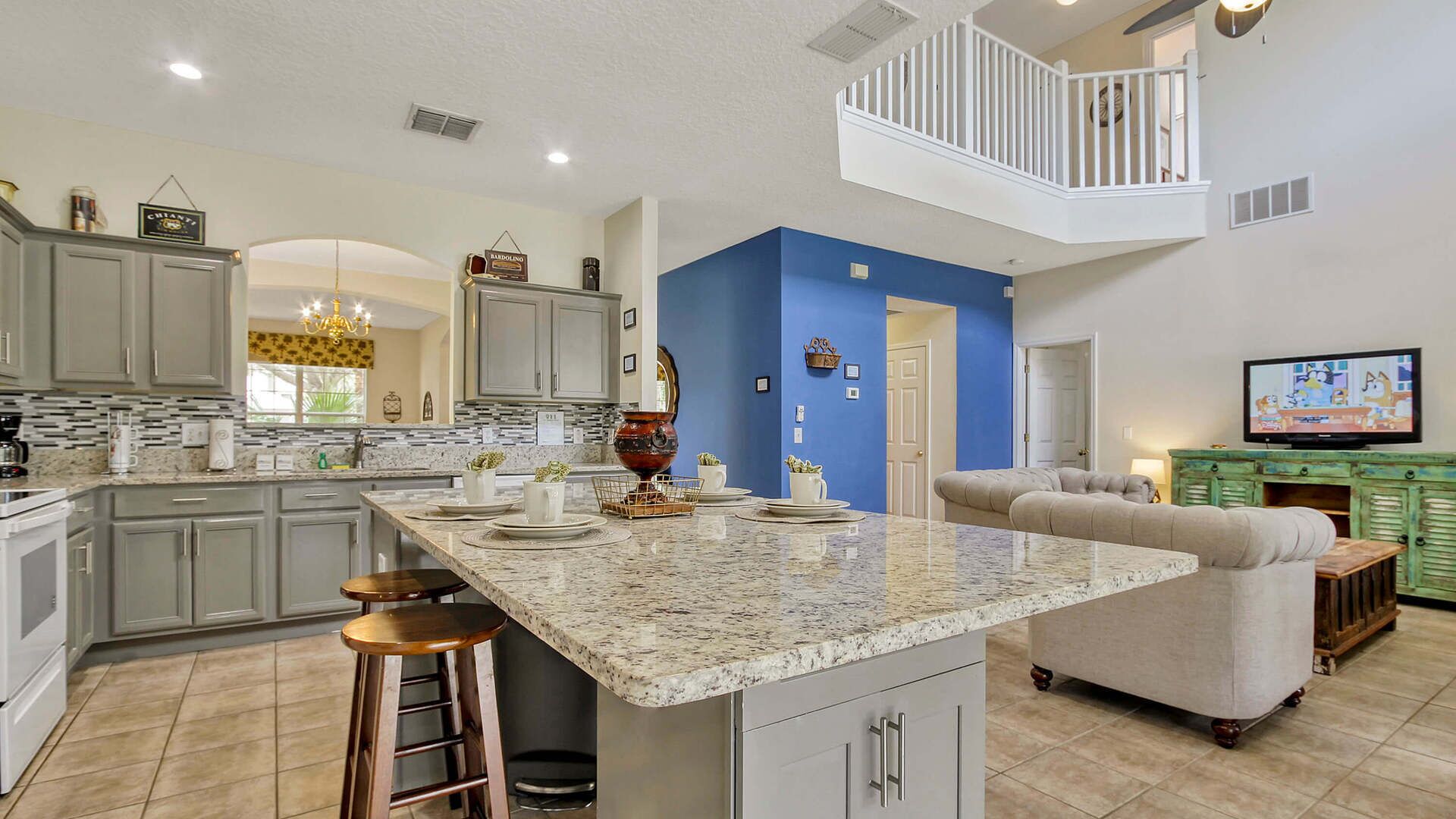 A modern kitchen with granite countertops, grey cabinets, and a central island with stools. A living area with a sofa and a television is visible in the background.