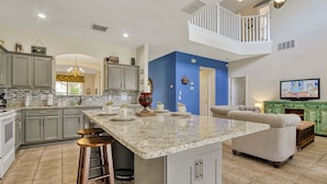 A modern kitchen with granite countertops, grey cabinets, and a central island with stools. A living area with a sofa and a television is visible in the background. - Spacious 6 bed pool home with spa and games room in Emerald Island, sleeps 15 (Kissimmee)