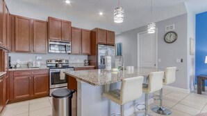 A modern kitchen with wooden cabinets, stainless steel appliances, a granite countertop, and a bar area with stools. - Enclave at Festival: Your Private Oasis of Luxury + Comfort - Davenport, Florida (Davenport)