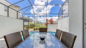 A covered outdoor area with a glass table and wicker chairs, overlooking a swimming pool and garden. - Enclave at Festival: Your Private Oasis of Luxury + Comfort - Davenport, Florida (Davenport)