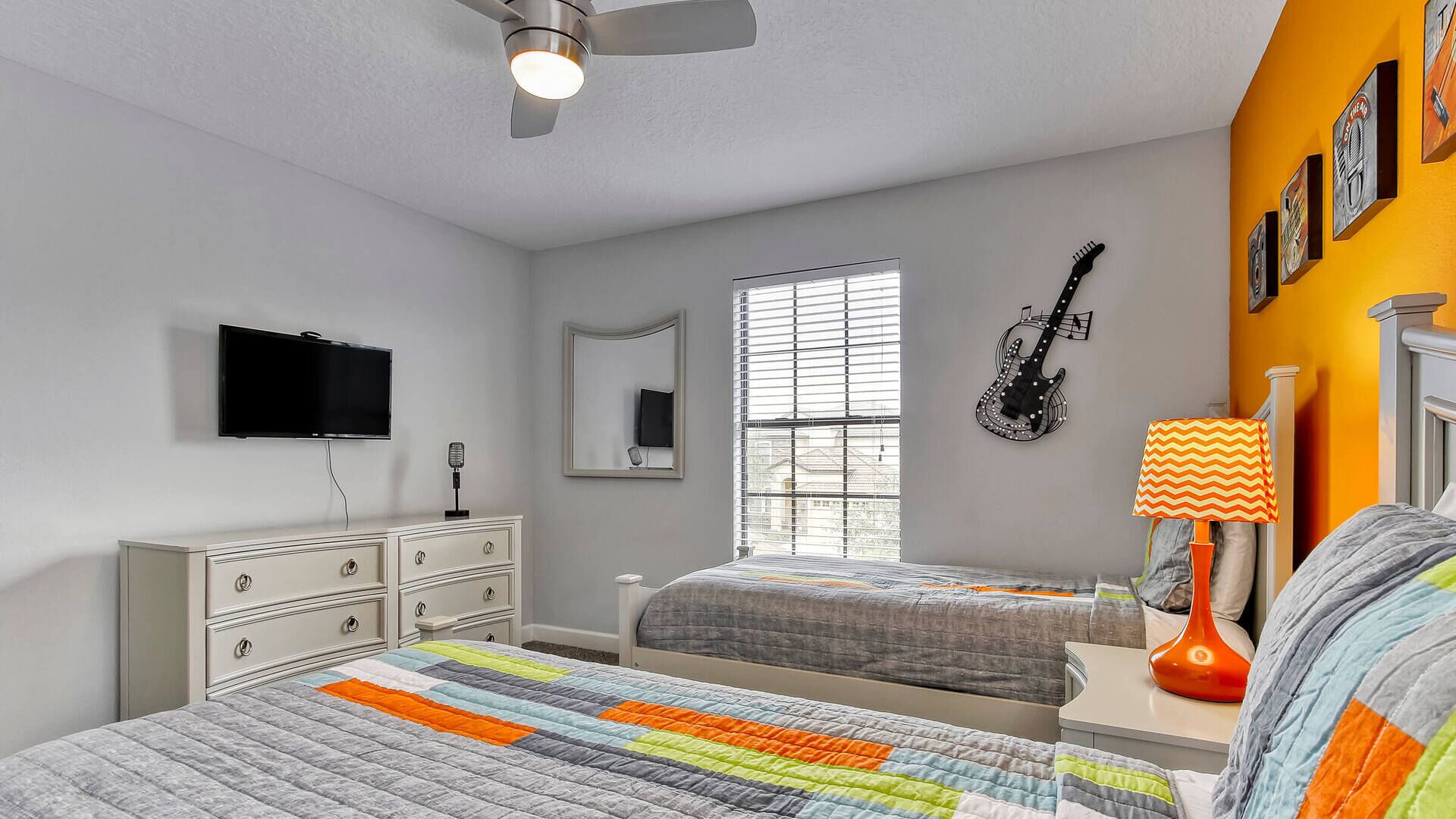 A bedroom with two beds, a ceiling fan, a TV, a guitar, and a window with blinds.