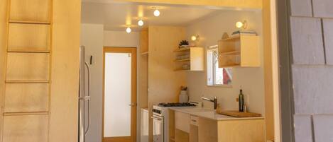 A compact kitchen with a refrigerator, stove, and white cabinets. A wooden ladder is against the wall, and a white chair is visible.