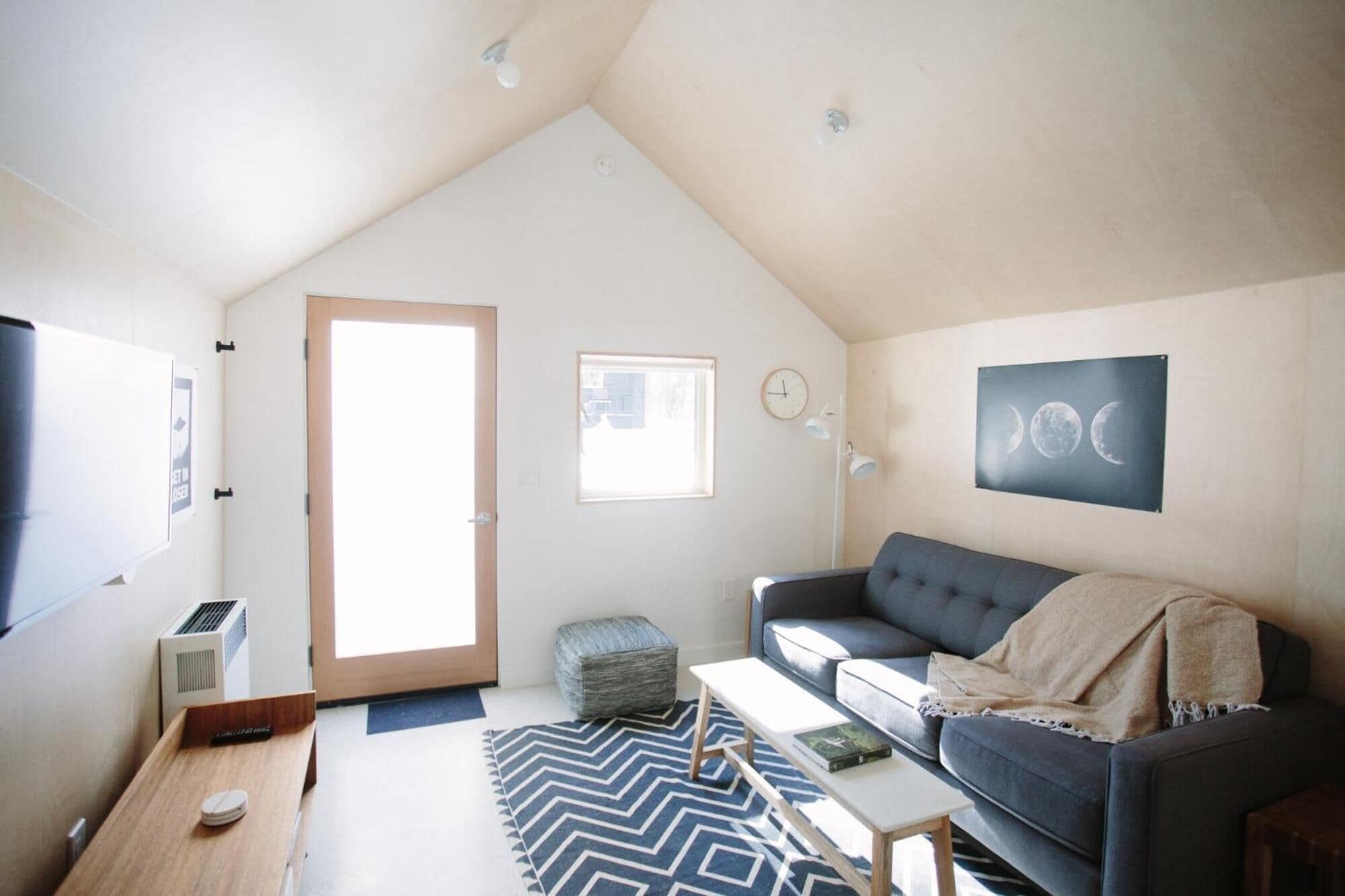 En hyggelig loftstue med højt til loftet, et stort vindue, en mørkegrå sofa, et lille bord og et vægmonteret ur.