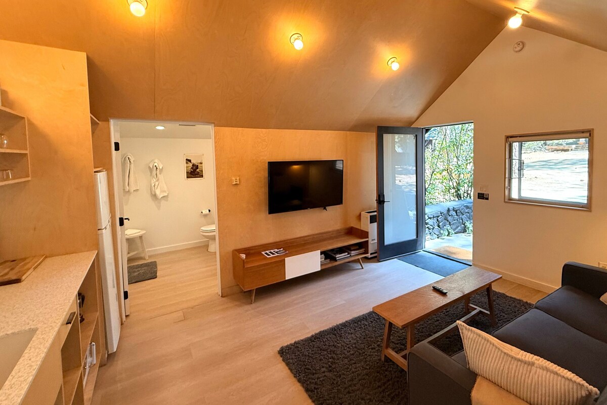 A modern living room with a flat-screen TV, a sofa, a coffee table, and a kitchen area with a refrigerator and a countertop.