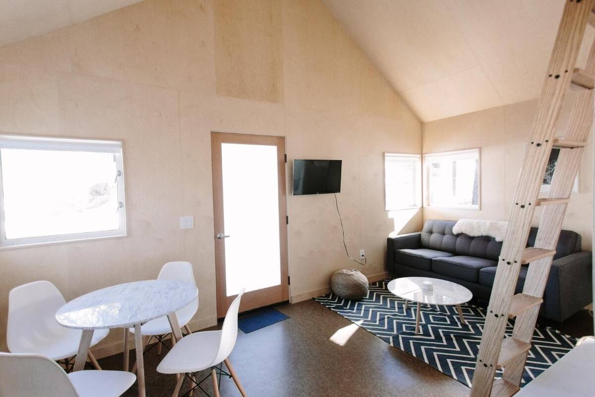 A spacious room with a high ceiling, a grey sofa, a white table with chairs, and a television mounted on the wall.