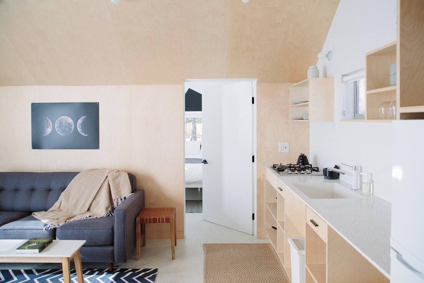 A modern living room with a sofa and a coffee table, a kitchen with a stove and cabinets, and a door leading to another room.