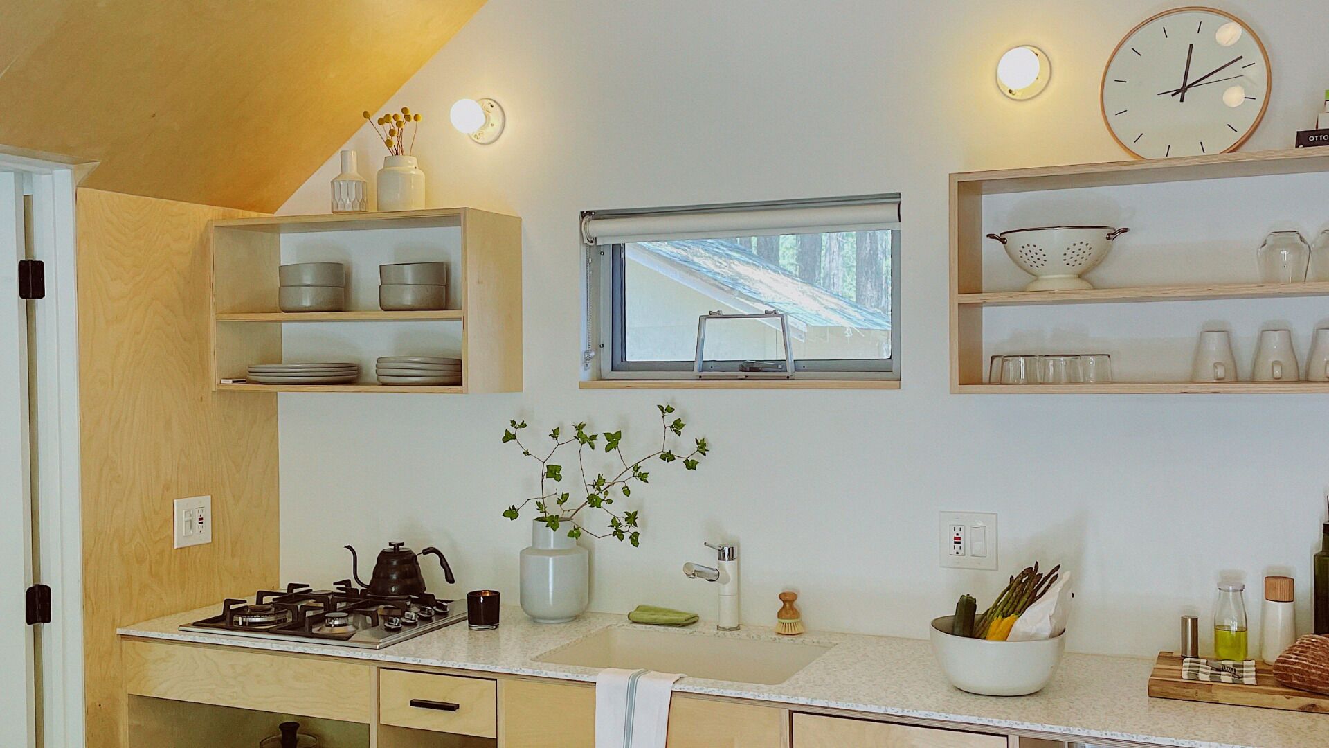 A kitchen with a stove, sink, and window. There are shelves with dishes and a clock on the wall.