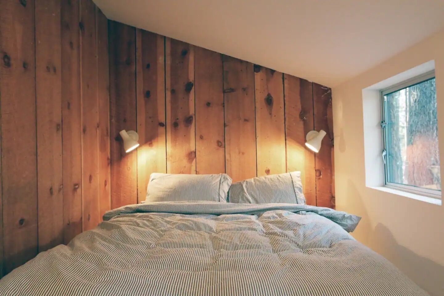 A bedroom with a wooden headboard, two bedside lamps, a bed with striped bedding, and a window with a view of trees.
