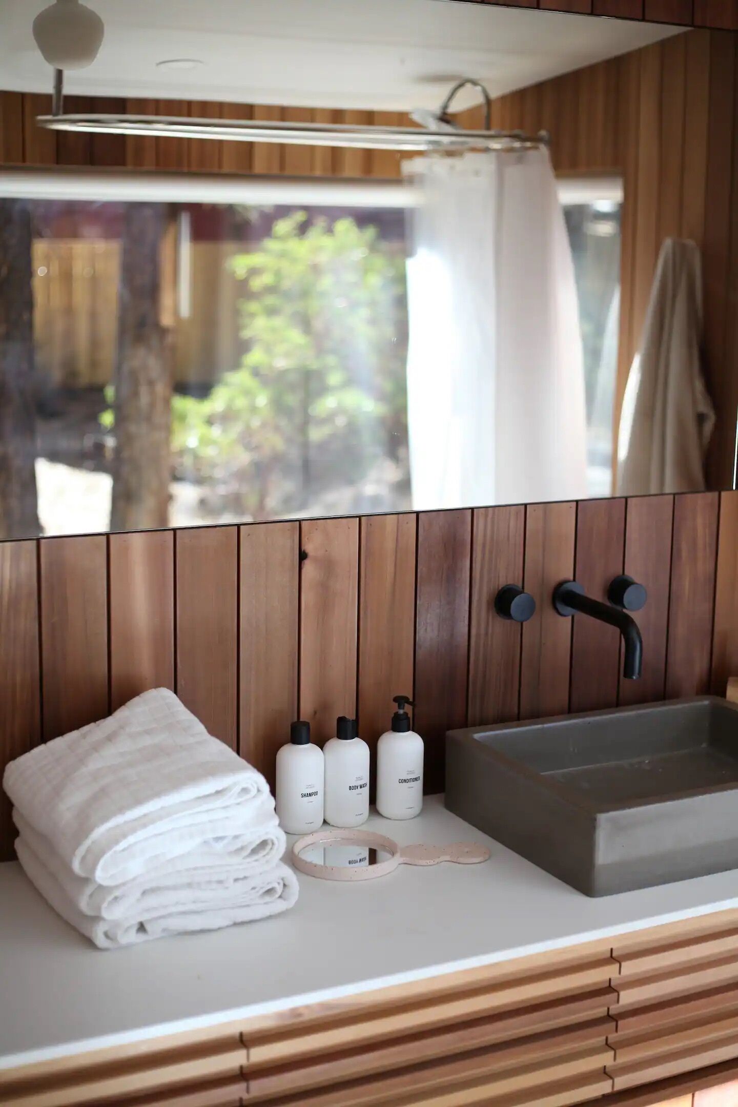 A bathroom with a wooden panel design, a sink with two faucets, and a mirror. There are towels stacked on the counter and three bottles of toiletries.