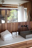 Un baño con paneles de madera, un lavamanos con dos grifos y un espejo. Hay toallas apiladas en la encimera y tres botellas de artículos de aseo.