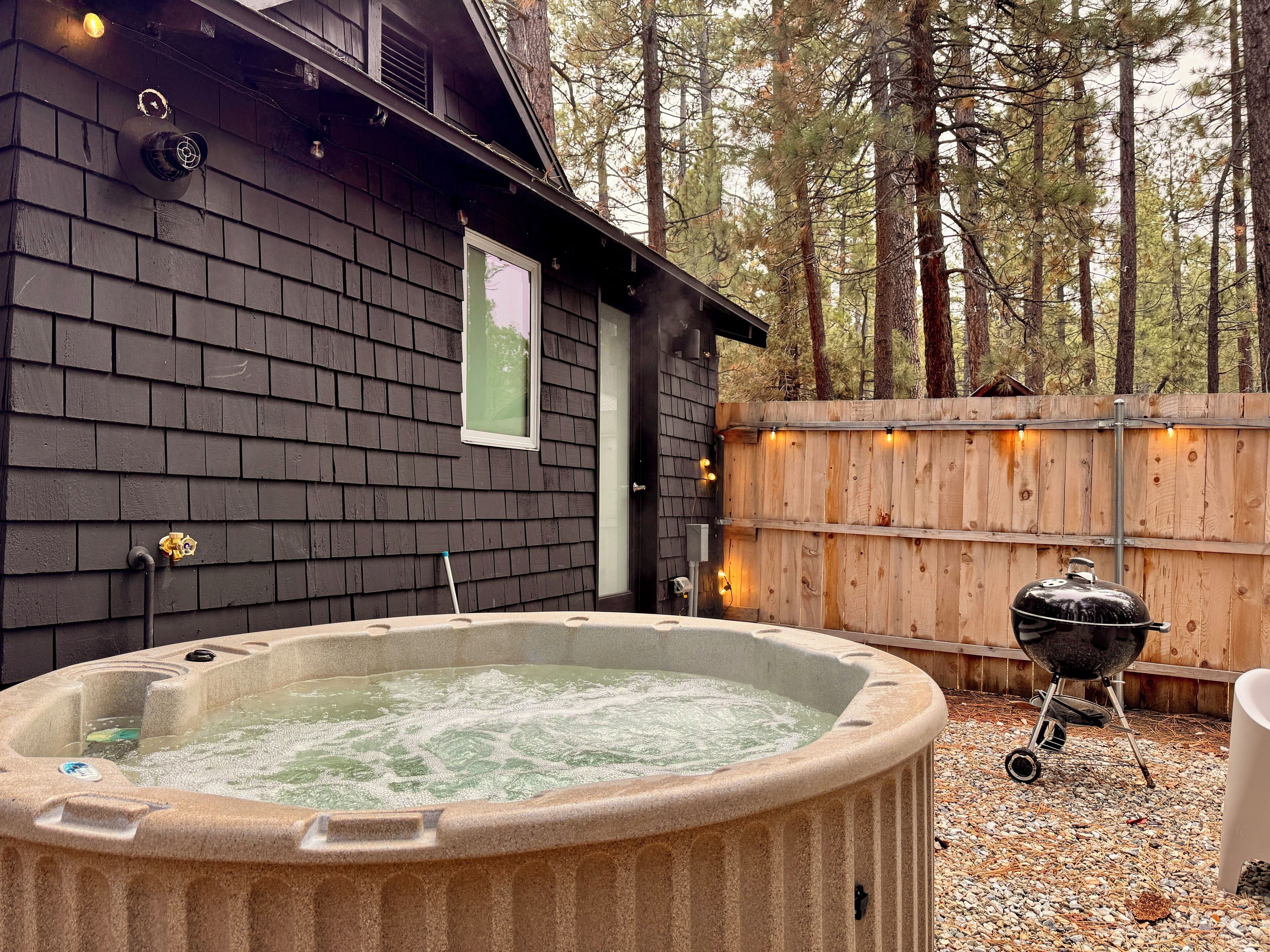 Un jacuzzi en un patio trasero con terraza de madera, una parrilla y una valla de madera.
