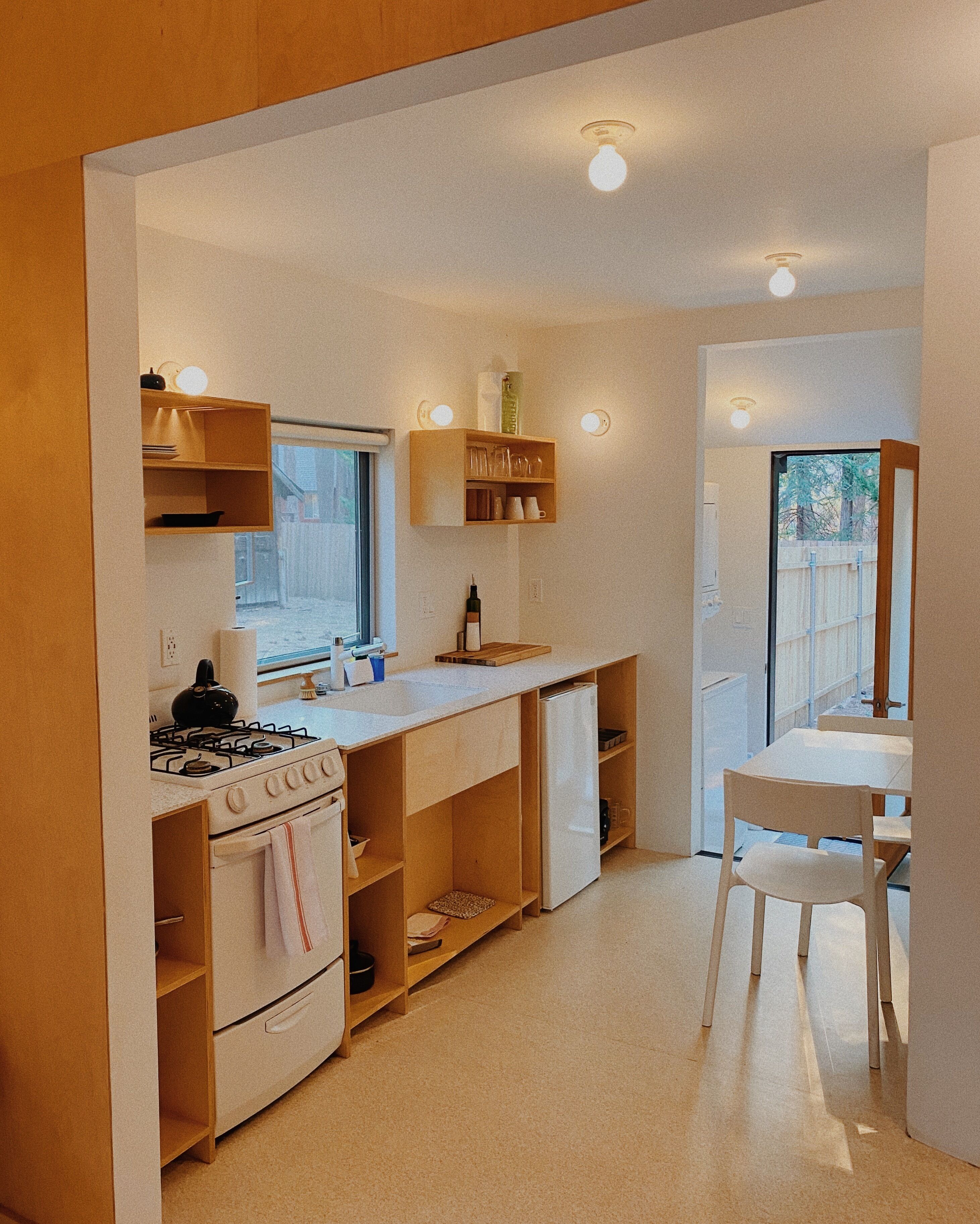 A modern kitchen with built-in appliances, wooden shelves, and a dining area with a white table and chair.