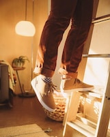 A person standing on a wooden ladder in a room with a lamp, a plant, and a rug.