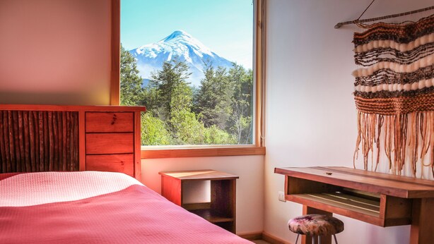 A bedroom with a bed, a wooden desk, a stool, and a view of a mountain through a window. - Hamilton's Place (Puerto Varas)