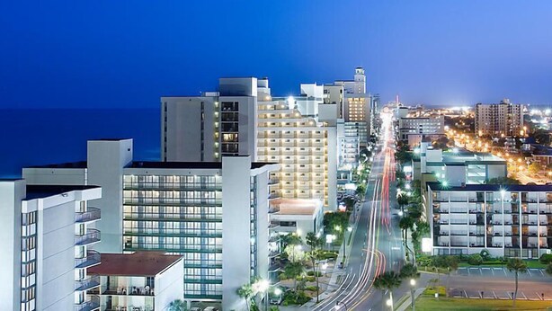 A modern hotel with multiple floors, a central courtyard, and a view of the cityscape at dusk. - Located in the Heart of Myrtle Beach, close to the Beach, Museum's, with access to all amenities (Myrtle Beach)