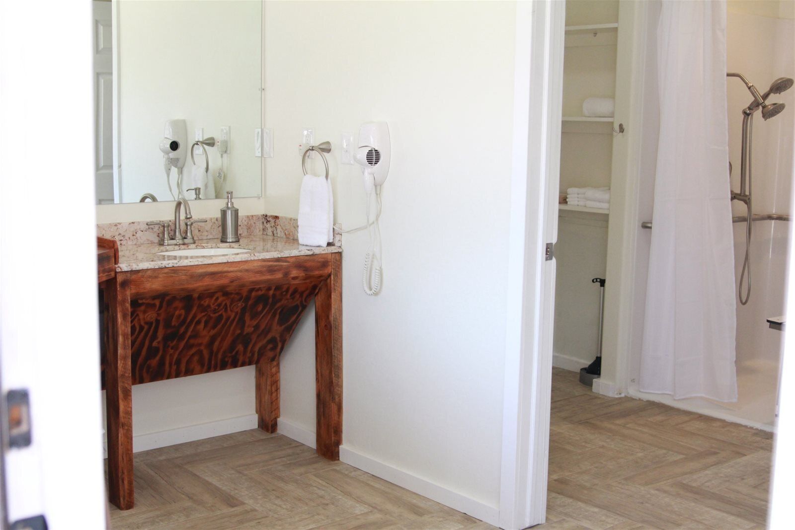 A bathroom with a wooden vanity, a mirror, a hairdryer, and a shower area.