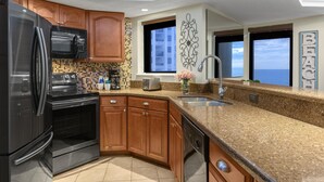 Una cocina moderna con refrigerador de acero inoxidable, microondas negro y horno, armarios de madera, encimera de granito y vistas al océano a través de amplias ventanas.