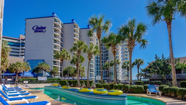 A pool area with lounge chairs, palm trees, and a hotel building with balconies. - Angle Oceanfront King Suite at Fun Resort + Official On-Site Rental Privileges (Myrtle Beach)