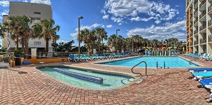 Zona de piscina de hotel con dos piscinas, sillas de descanso, palmeras y un paseo de ladrillo.